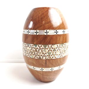 May include: An oval-shaped wooden vase. The vase is crafted from brown wood and adorned with three bands of decorative inlay. The top and bottom bands feature a white background with black cross-shaped patterns. The central band displays a geometric design.