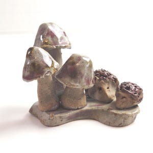 May include: A ceramic sculpture of three mushrooms with burgundy caps and two small hedgehog figures. The mushrooms are on a grey base. The sculpture has a rustic, handmade appearance.