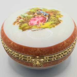 May include: A round porcelain trinket box with a gold trim and a floral design. The lid features a painted scene of a couple in a garden setting.