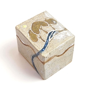 May include: A small, rectangular ceramic box with a lid. The box is beige with a tree and river design in brown and blue. A yellow circle represents the sun. The box is likely used for storing small items.