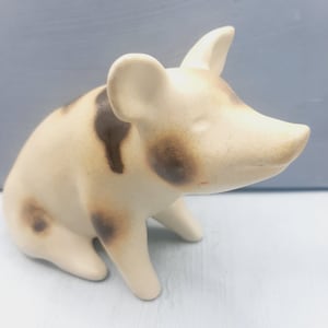 May include: A ceramic pig figurine with brown spots. The pig is sitting with its head tilted to the side.