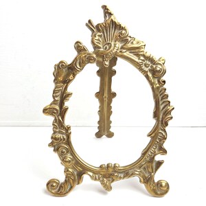 May include: An antique-style brass picture frame with an oval aperture. The frame has elaborate floral and scrollwork patterns, supported by a decorative stand. The frame is a warm, golden colour, and is set against a white background.