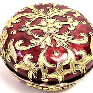 May include: A round trinket box with a deep red enamel finish and gold-coloured filigree. The box has a floral design and is likely made of metal. The box is a decorative item.