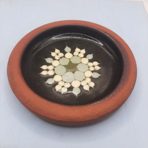 May include: A terracotta dish with a black glaze and a white and green floral design in the centre.