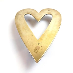 May include: A heart-shaped object in a brass colour with a hollow centre. The metal has a slightly aged appearance with minor surface imperfections. The heart's edges are smooth and rounded, set against a white background.