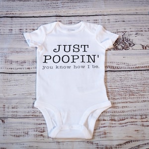 May include: White baby onesie with the text "JUST POOPIN' you know how I be." in black. The onesie is centered on a light-colored, wooden surface.