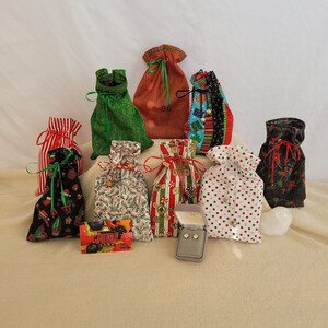 May include: A collection of nine small fabric gift bags with various Christmas-themed patterns. The bags are filled with small items and are arranged in a pyramid shape. A small box with a pair of earrings is in the center of the image.