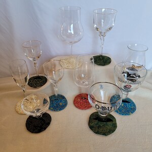 May include: A collection of nine clear glass drinking glasses with gold rims, each sitting on a colorful patterned coaster. The coasters feature various designs, including swirls, floral patterns, and geometric shapes.
