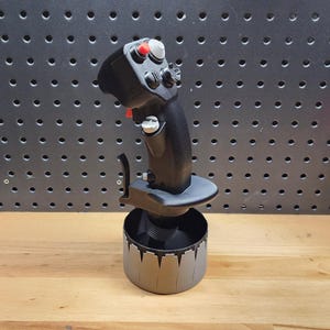 May include: A black joystick with red and gray buttons and a gray base. The joystick has a curved handle and several buttons and switches. The base is cylindrical with a decorative edge. The background is a dark gray pegboard.