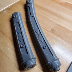 May include: Two gray plastic lightsaber hilts with diamond-shaped patterns. The hilts are curved and have a textured surface.