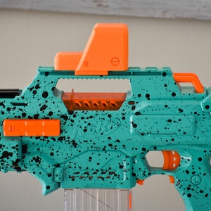 May include: A teal and black Nerf blaster with an orange scope and trigger. The blaster has a black and white splatter paint design.