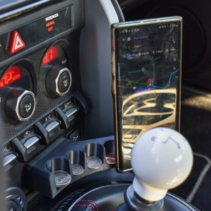 May include: A black car interior with a phone holder mounted to the center console. The phone is displaying a map app with a red pin indicating the current location. The phone is mounted next to a white gear shift knob.