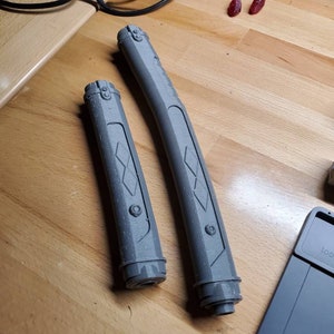 May include: Two gray plastic lightsaber hilts with a diamond pattern and a circular detail. The hilts are on a wooden surface.