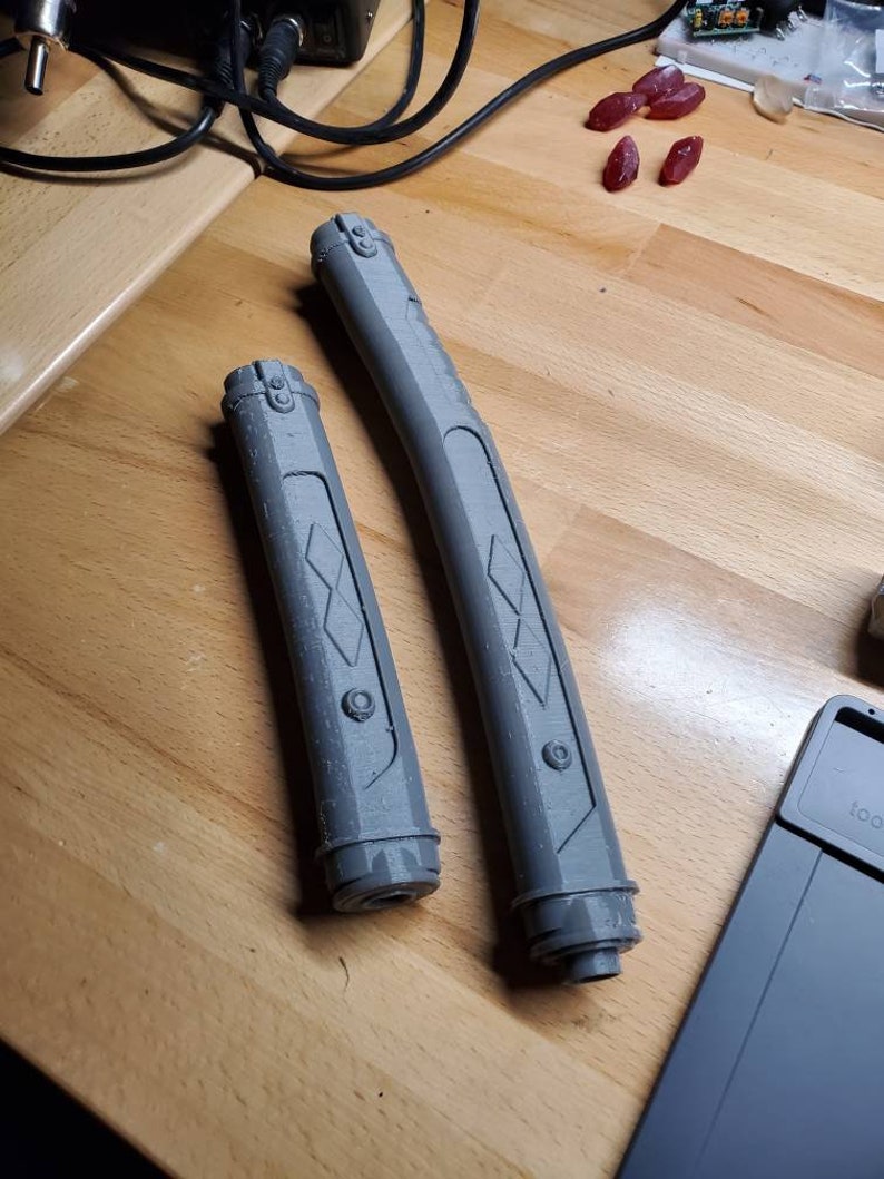 May include: Two gray plastic lightsaber hilts with a diamond pattern and a circular detail. The hilts are on a wooden surface.