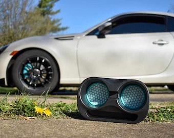 May include: Black car audio speaker with two circular openings featuring a honeycomb pattern and blue accents. The speaker is positioned on a concrete surface with a white car in the background.