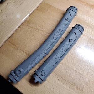 May include: Two gray plastic lightsaber hilts with diamond-shaped patterns and a textured grip. The hilts are on a wooden surface.