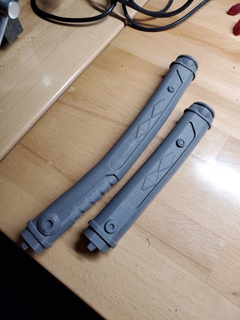 May include: Two gray plastic lightsaber hilts with diamond-shaped patterns and a textured grip. The hilts are on a wooden surface.