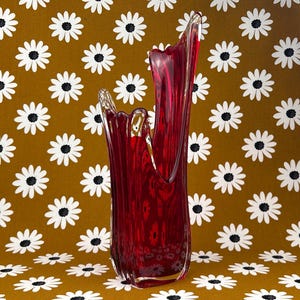 May include: A tall, deep red glass vase with an asymmetrical, undulating rim. The vessel features a smooth, translucent surface with vertical ribbing that catches the light, creating varying shades of crimson throughout the structure.
