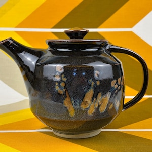 May include: A dark brown ceramic teapot with a glossy finish, featuring a wide body, a curved handle, a pointed spout, and a matching lid with a small knob. The surface displays mottled tan and blue glaze accents across the rounded exterior.