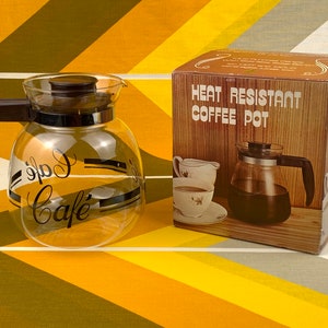 May include: A clear glass coffee pot with a black handle and a black "Cafe" logo. The pot is sitting on a yellow and brown striped surface. The box for the coffee pot is in the background and says "Heat Resistant Coffee Pot".