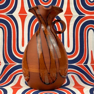 May include: A brown ceramic jug with a textured, striped design and a handle. The jug has a wide mouth and a rounded body.