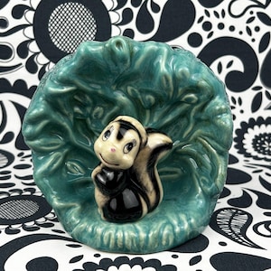 May include: A vintage ceramic skunk ornament in a teal leaf-shaped planter. The skunk has a black and white coat and a pink nose. The planter has a textured surface. The background is a black and white patterned fabric.