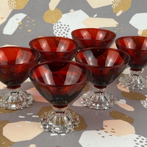 May include: A set of six vintage ruby red glass dessert bowls. Each bowl has a clear, textured pedestal base. The bowls are displayed on a patterned grey and gold surface. The bowls are suitable for serving desserts.