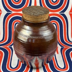 May include: A brown ceramic jar with a cork lid. The jar has a decorative band of small circles around the middle. The jar is sitting on a white and red patterned fabric.