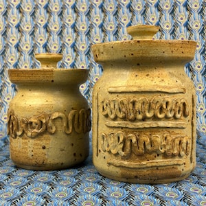 May include: Two tan ceramic canisters with lids, featuring speckled textures and raised, wavy clay detailing around the bodies. The smaller canister has one row of wavy decoration, while the larger canister features two rows of wavy clay lines.