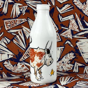May include: A white ceramic milk bottle featuring a cartoon cow with brown markings. The cow faces right, with a small yellow chick beside it. The bottle has a narrow neck and a rounded body, suitable for approximately 1 litre.