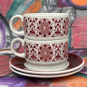 May include: Two white ceramic teacups with a red floral pattern stacked on top of each other. The cups are sitting on a matching saucer.