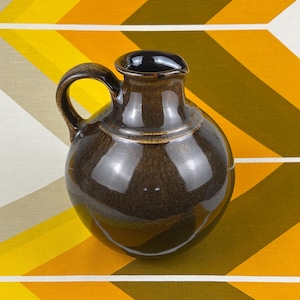 May include: A round, dark brown ceramic pitcher with a glossy, reflective glaze. The vessel features a short, narrow neck, a wide, bulbous body, and a single curved handle attached to the side. The interior of the rim is dark and smooth.