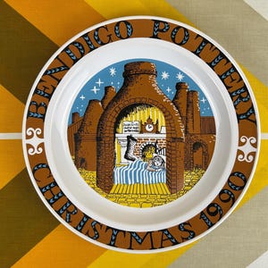 May include: A white ceramic plate with a brown and blue border. The plate features a Christmas scene with a brick building, a fireplace, and a sleeping child. The text "Indigo Pottery Christmas 1990" is printed around the border.