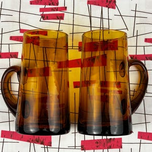 以下が含まれることがあります： アンバー色のガラス製ビールジョッキ2個。各ジョッキは、赤い長方形と黒い線のデザインが特徴です。ジョッキは、赤と黒のパターンを持つ白い背景に配置されています。