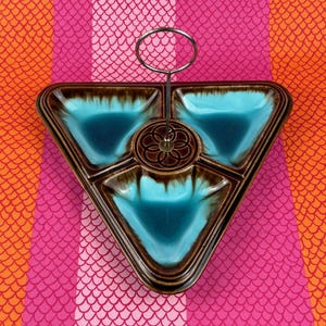 May include: A triangular ceramic serving dish with three individual compartments. The dish is brown with a blue glaze and has a metal stand in the center.