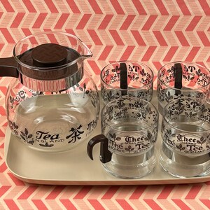 May include: A retro tea set comprising a glass teapot and four mugs, each adorned with black floral and text patterns. The teapot and mugs feature brown handles and lids. The set is presented on a beige tray, set against a pink and white patterned backdrop.
