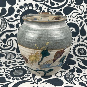 May include: A ceramic jar with a lid. The upper section is grey, the lower section is cream-coloured, and it has colourful paint splatters in blue, yellow, and brown. This decorative jar would suit any home.