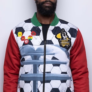Może przedstawiać: Biała kurtka bomber z wzorem piłki nożnej i czerwonymi i zielonymi rękawami. Kurtka ma zamek błyskawiczny i naszywkę na piersi z napisem "Forward Ever Backward Never".