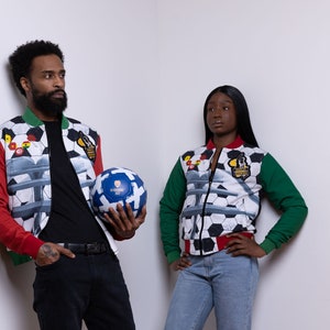 May include: Two people wearing red, green, and white patterned jackets with a soccer ball design. The jackets have a zipper closure and a black collar. The person on the left is holding a soccer ball.