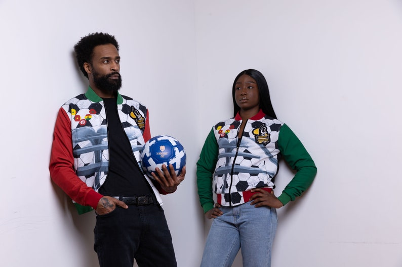 May include: Two people wearing red, green, and white patterned jackets with a soccer ball design. The jackets have a zipper closure and a black collar. The person on the left is holding a soccer ball.