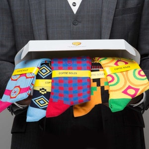 May include: A white box with the text "Coptic Soles" on it. The box is open and contains five pairs of colorful socks with different patterns. The socks are folded and arranged in a row.