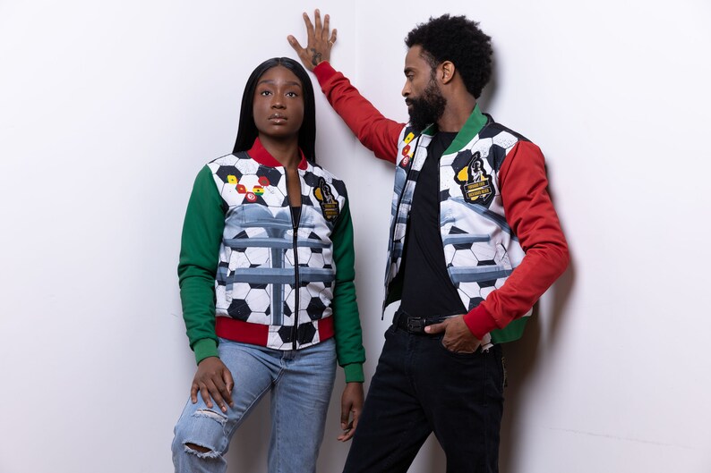 May include: Two people wearing red, green, and white patterned jackets with a soccer ball design. The jackets have a zipper closure and a black background with white soccer balls and a gray design. The jackets also have a green and red sleeve design.