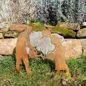 Può includere: Due sagome metalliche arrugginite di persone che annaffiano le piante. Le figure sono in piedi su un letto di erba verde davanti a un muro di pietra.