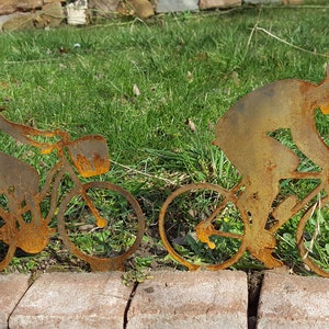 Könnte beinhalten: Zwei rostige Metallsilhouetten von Personen, die Fahrrad fahren. Die Figuren sind nach rechts gerichtet und vor einem grünen grasbewachsenen Hintergrund platziert.
