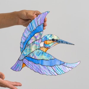 K&ouml;nnte beinhalten: Ein Buntglasvogel im Flug, in Blau-, Lila- und Oranget&ouml;nen. Die Fl&uuml;gel des Vogels sind ausgebreitet und zeigen detaillierte Muster und schillernde Effekte. Das Kunstwerk wird von einer Hand gehalten, was die Handwerkskunst hervorhebt.