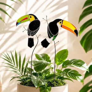 May include: Two stained glass toucan plant stakes featuring black bodies, white accents, and vibrant beaks in lime green and bright orange. Each bird is mounted on a thin, dark metal rod, designed to be inserted into a potted plant for decoration.
