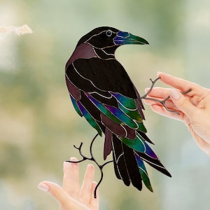 Puede incluir: Un vitral de un cuervo negro con detalles morados y plumas iridiscentes verdes y azules. El cuervo está posado en una delicada rama de metal oscuro. El pico es verde. La obra de arte es sostenida por una persona.