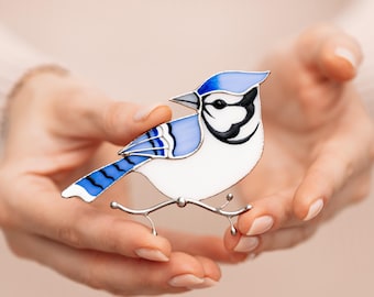 Pode incluir: Broche de pássaro gaio-azul com corpo branco, asas azuis e cabeça preta e branca. O pássaro está empoleirado em um galho prateado. O broche é segurado nas mãos de uma pessoa.