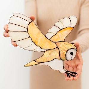 Könnte beinhalten: Eine Scheunen-Eule aus Buntglas mit weißen und gelben Flügeln und schwarzen Augen. Die Eule ist im Flug mit ausgebreiteten Flügeln.