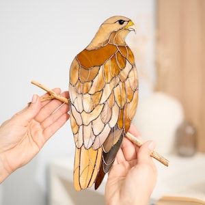 Puede incluir: Una escultura de halcón de vidrieras en tonos marrones, beige y crema, posada en una rama de madera. Las plumas del ave están meticulosamente elaboradas con un diseño en capas. El halcón tiene un pico amarillo y está sostenido por dos manos.